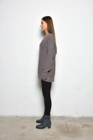 A woman standing against a white wall, wearing a 2 tone gray long-sleeve sweater with asymmetrical hem and black leggings.