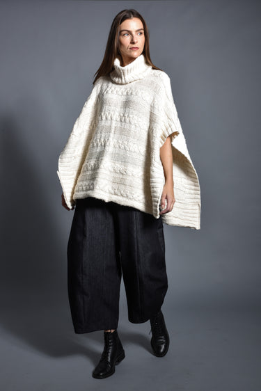 Woman wearing a cream knit turtleneck poncho with wide-leg black pants and black boots