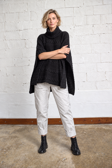 A woman stands against a white painted brick wall wearing a black ribbed turtleneck poncho-style sweater, light beige cropped pants, and black lace-up ankle boots.