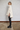 A woman stands in profile wearing a chunky cream knit turtleneck poncho over a black top and black ankle boots, posed against a white painted brick wall.