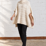 A woman modeling a chunky knit cream-colored poncho sweater, paired with black pants and black leather boots, standing in front of a white brick wall.