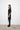 Woman wearing a black dress and black boots standing against a white wall.