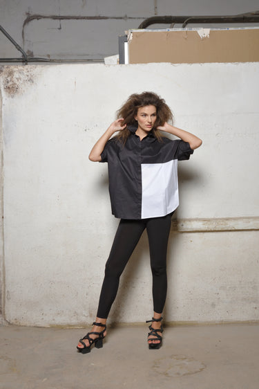 A woman modeling a 2 tone black and white boxy shirt with dolman sleeves, paired with black leggings and strappy sandals, standing against a textured wall.
