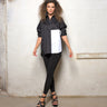 A woman modeling a 2 tone black and white boxy shirt with dolman sleeves, paired with black leggings and strappy sandals, standing against a textured wall.
