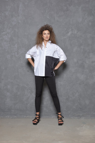 A woman modeling a 2 tone white and black boxy shirt with dolman sleeves, paired with black leggings and strappy sandals, standing against a textured wall.