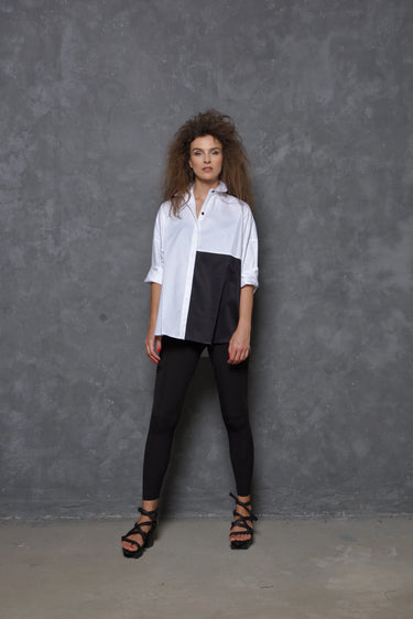 A woman modeling a 2 tone white and black boxy shirt with dolman sleeves, paired with black leggings and strappy sandals, standing against a textured wall.