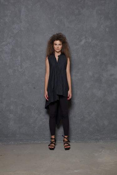 A woman modeling a sleeveless asymmetrical tunic shirt in black, paired with black pants and black strappy sandals, standing against a gray background.