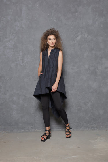 A woman modeling a sleeveless asymmetrical tunic shirt in black, paired with black pants and black strappy sandals, standing against a gray background.