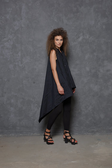 A woman modeling a sleeveless asymmetrical tunic shirt in black, paired with black pants and black strappy sandals, standing against a gray background.