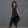 A woman modeling a sleeveless asymmetrical tunic shirt in black, paired with black pants and black strappy sandals, standing against a gray background.